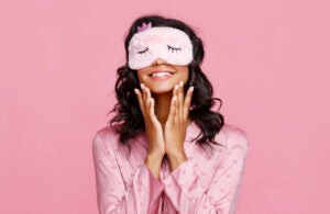 Delighted ethnic female in pajama and mask smiling and smearing lotion on face during skin care routine before sleep against pink background