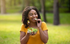 Pretty black girl eating fresh vegetable salad and winking at green park on summer day. Panorama