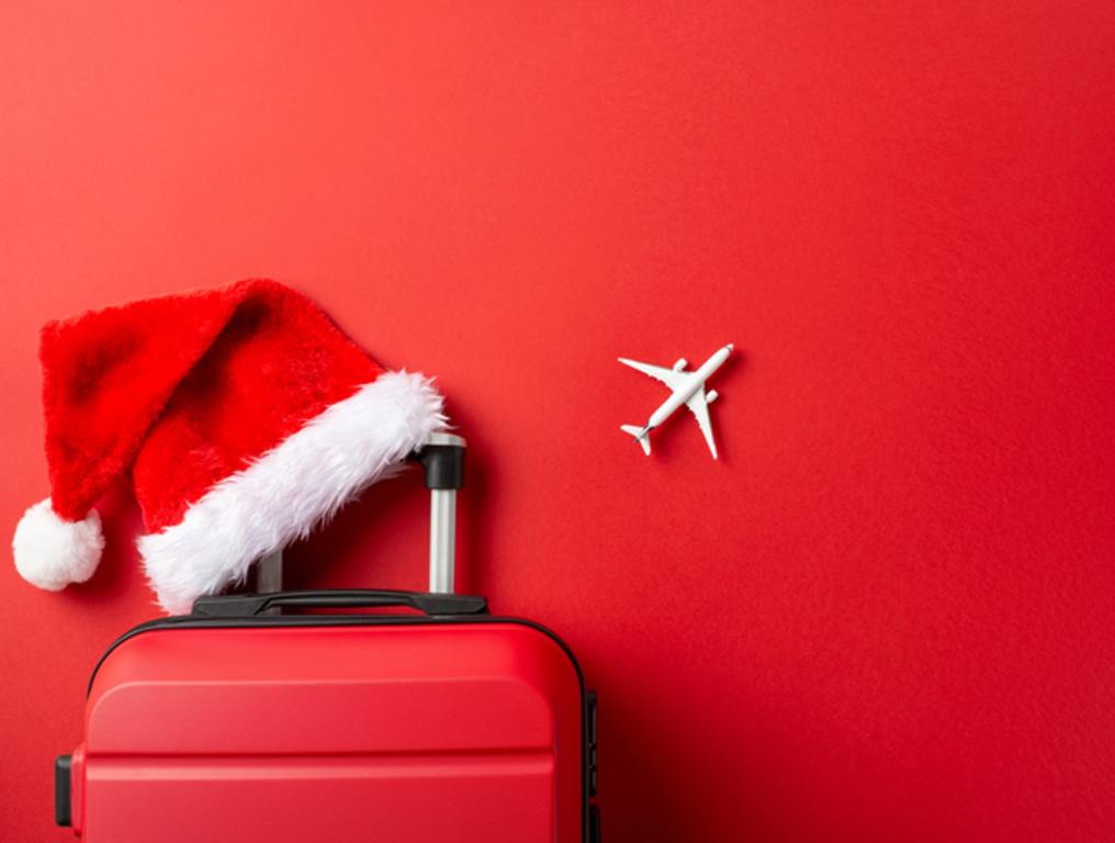 Top-view image of a suitcase, miniature airplane model, and Santa's hat on a vibrant red backdrop, holiday travel concept