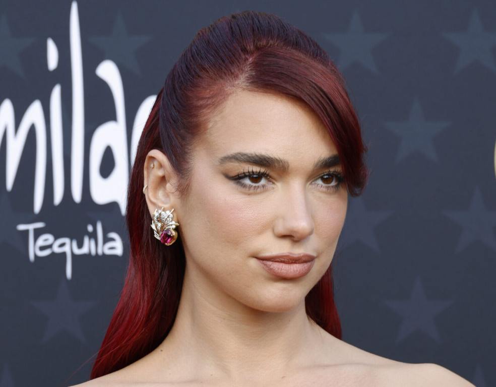 Dua Lipa attends the 29th Annual Critics Choice Awards facing right smirking with red hair.