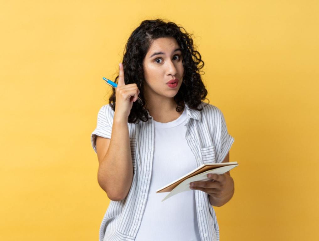 Portrait of amazed inspired woman journalist with dark wavy hair writing in paper notebook, having new excellent idea, raising finger up. Indoor studio shot isolated on yellow background.