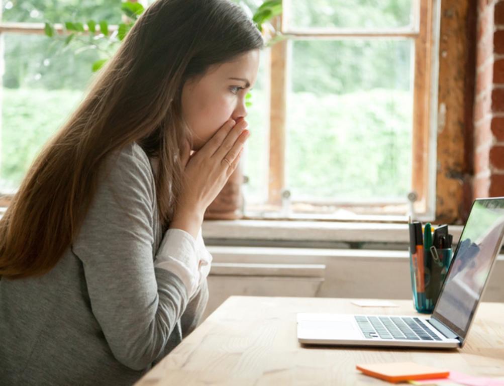 Shocked young woman looking at laptop screen at work desk. Casual business lady in office seeing something unbelievable on computer. Rejection concept.