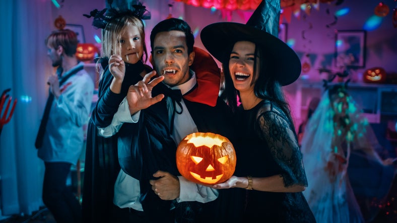 Halloween Costume Party: Father Count Dracula Holds Little Bat Girl Daughter and Hugs Witch Wife for a Happy Family Portrait. In the Background Monsters Having Fun. Popular 2024 Halloween Costumes