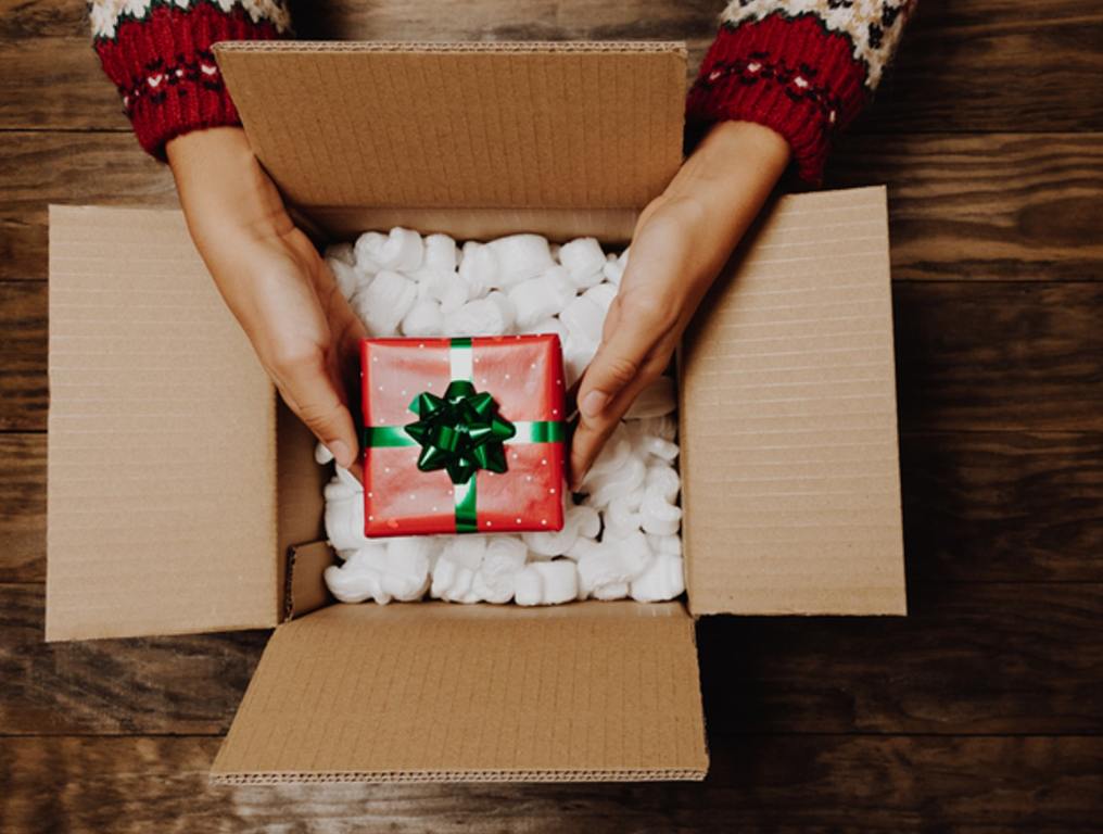 Female hands with a winter sweater packing or unpacking a Christmas gift from a cardboard box on a rustic wooden table. Christmas home delivery or shipping concept.